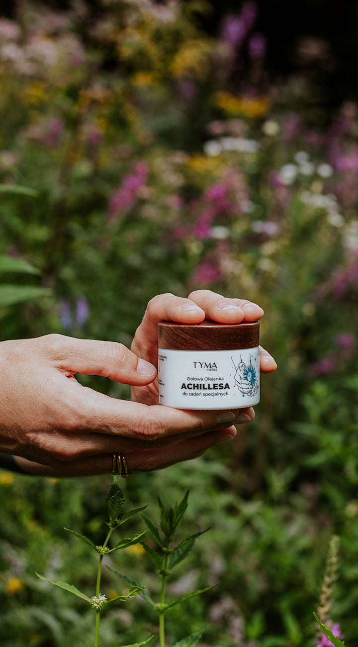 Olejanka Achillesa Tyma Herbs trzymana w dłoniach na tle dzikiego ogrodu ziół – naturalny krem na AZS